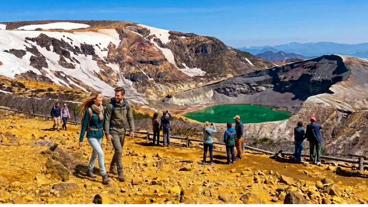 Hikers on the yellow volcanic rim above the green Okama crater lake on Mount Zao, snow still on the slopes