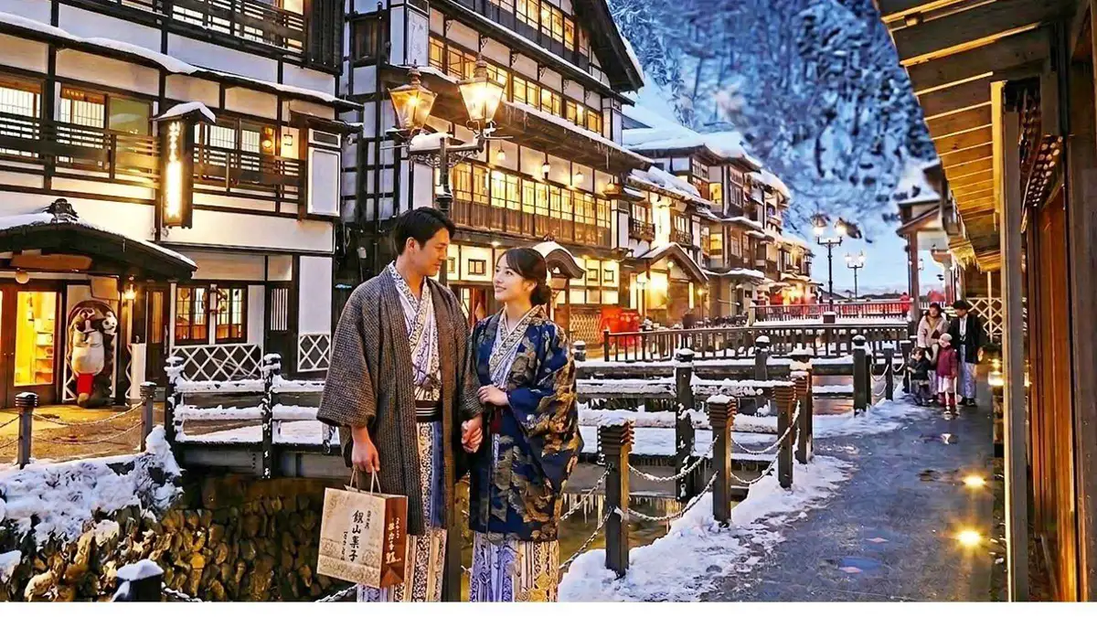 A couple in yukata walking along the snowy riverside street of Ginzan Onsen at dusk with gas lamps lit