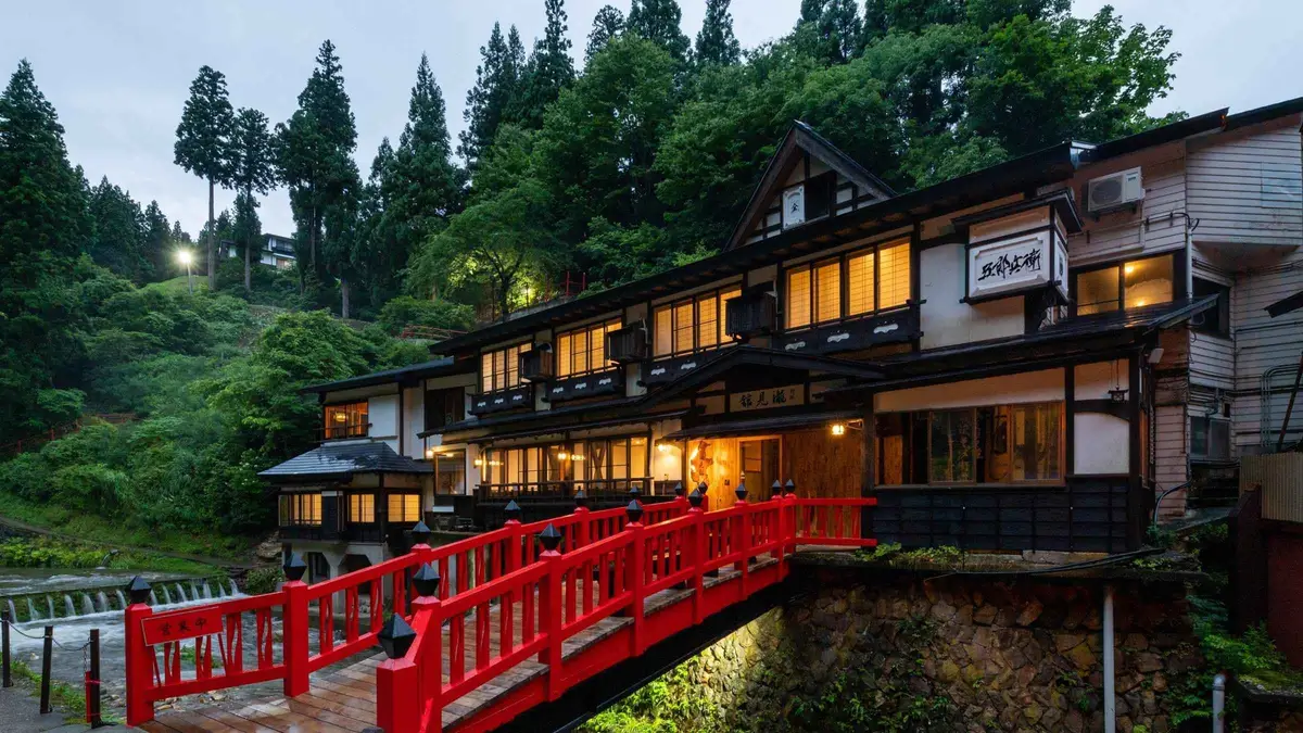 A red wooden bridge in front of a lantern-lit ryokan at Ginzan Onsen under cedar forest