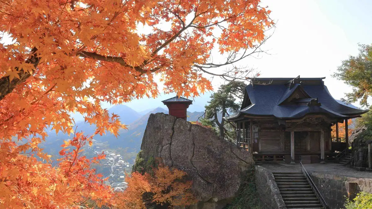 A temple hall on a rocky outcrop at Yamadera framed by bright orange maple leaves in autumn