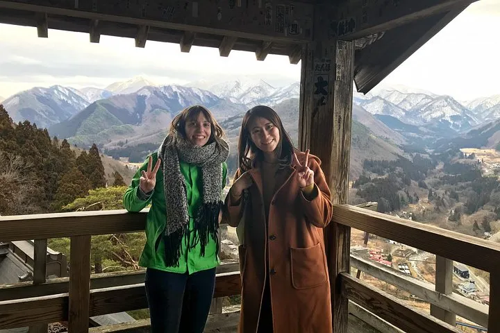 Two travellers on the wooden Godaido balcony at Yamadera with snow-dusted mountains behind them