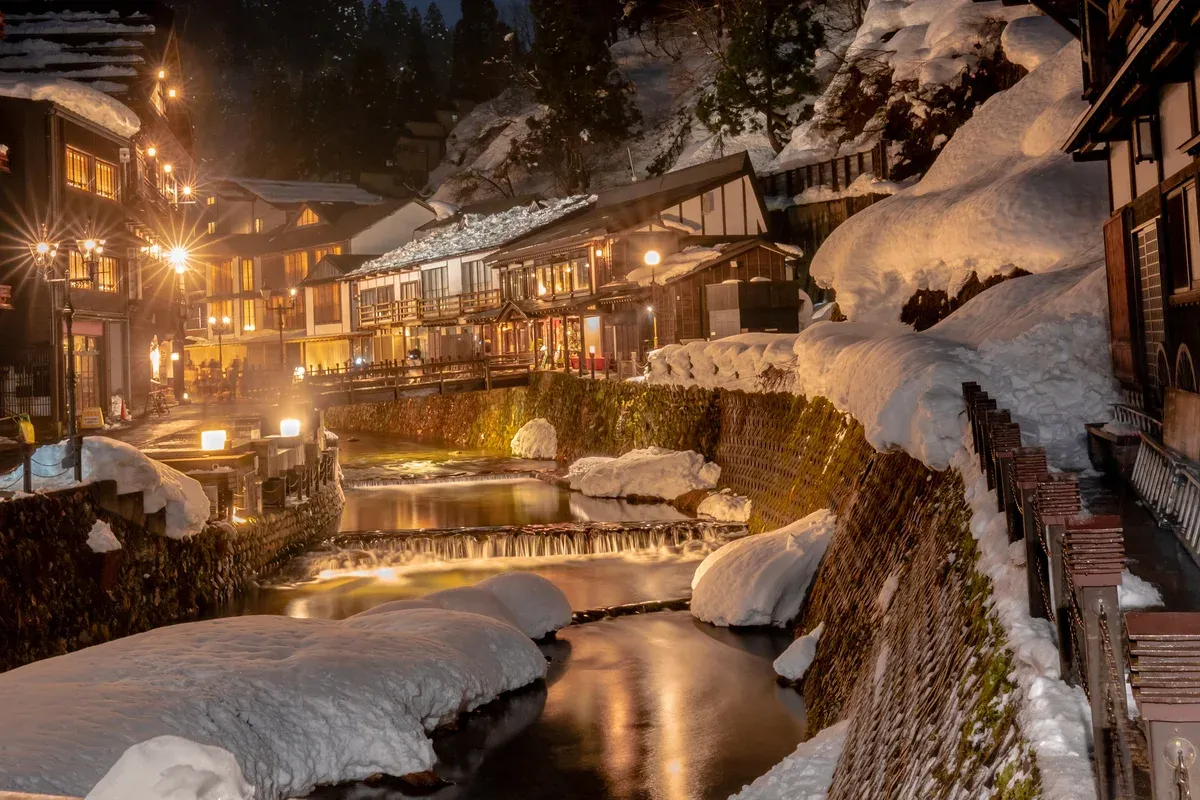 Snow piled along the river through Ginzan Onsen at dusk with lit wooden inns on both banks