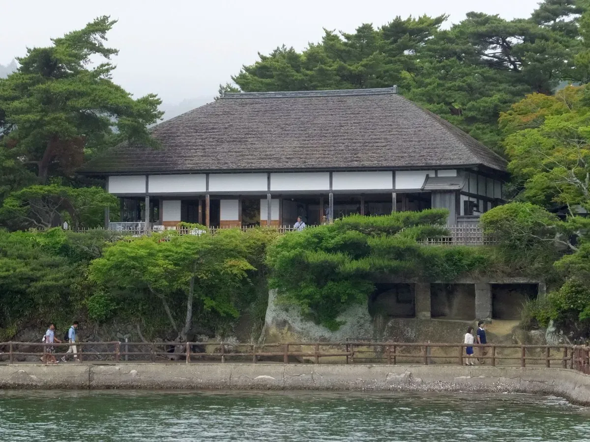 Thatched tea house on the rocky shore seen from the water, backed by pines